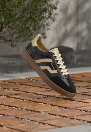 Zwarte en beige leren sneaker met beige veters en gumzool, getoond boven een houten tegelvloer met groen en een betonnen muur als achtergrond.