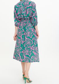 Mujer que lleva un vestido midi verde con estampado floral en rosa, mangas tres cuartos y sandalias de tacón verde, vista desde la espalda.