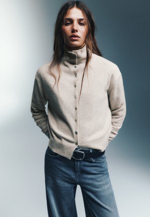 Une femme porte un cardigan crème boutonné à col montant, un jean foncé et une ceinture noire devant un fond de studio gris.