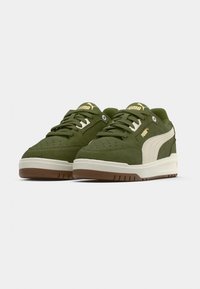 Ζευγάρι χαμηλά πράσινα σουέντ αθλητικά παπούτσια Puma με λευκές λεπτομέρειες και σόλες σε χρώμα καραμέλας, τοποθετημένα δίπλα δίπλα σε ουδέτερο φόντο.