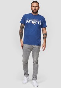 Sininen puuvillapaita, jossa on teksti "New England Patriots", lyhyet hihat, yhdistetty vaaleanharmaisiin farkkuihin ja valkoisiin lenkkareihin.