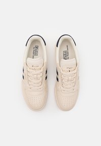 Een paar beige Polo Ralph Lauren sneakers met marineblauwe strepen, veters gestrikt en geperforeerde neuskapjes op een witte achtergrond.