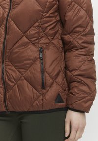 Veste matelassée brune avec des accents noirs, poche zippée et un écusson en forme de triangle à l'ourlet. Matériau texturé et résistant à l'eau.