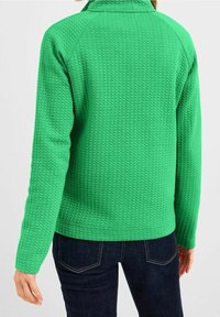 Chaqueta tipo pulóver texturizada verde con un patrón en relieve, que cuenta con cuello y mangas largas, combinada con jeans azul oscuro.