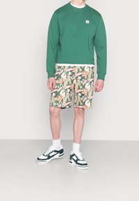 Sudadera verde con un pequeño logo, combinada con pantalones cortos florales que presentan patrones en rosa y verde, y zapatillas verdes y blancas.