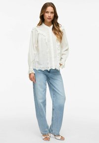 Biała koszula z guzikami, z falbanami na przodzie i mankietach, zestawiona z jasnoniebieskimi spodniami z wysokim stanem i białymi sandałami na obcasie.