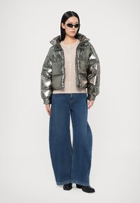 Metallic silver puffer jacket with large quilted sections, layered over a beige button-up sweater and paired with wide-leg denim jeans.
