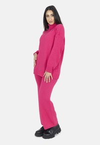 Heller pink gestrickter Pullover mit hohem Kragen und lockerer Passform, kombiniert mit passenden rosa Hosen. Schwarze klobige Schnürstiefel runden das Outfit ab.