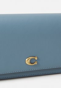 Portafoglio blu in pelle con texture liscia, dotato di un prominente logo dorato a forma di "C" e cuciture decorative lungo i bordi.