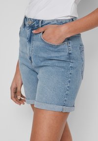 Calções de denim azul claro com cintura alta, bainhas enroladas, bolsos e fecho de botão. O tecido apresenta um ligeiro desbotado e uma textura suave.