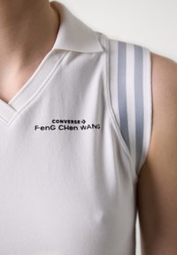 Polo shirt sans manches blanc avec un col en V, présentant des rayures grises sur les épaules et un logo imprimé "CONVERSE Feng Chen WANG" sur le devant.