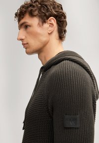 Donkergroene, gestructureerde hoodie met een trekkoordcapuchon en een vierkant logo-op-patch op de mouw, met een getailleerde pasvorm en geribbelde details.