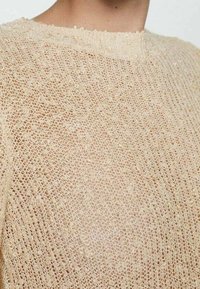 Beżowy sweter z dzianiny o luźnej, otwartej strukturze i subtelnym, falbaniastym dekolcie, wykonany z lekkiego i przewiewnego materiału.