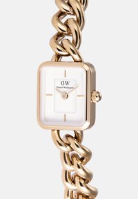 Kuldne kell, millel on ruudukujuline valge all dial. Omab kuldseid aktsente ja käsi, mille peal on musta teksti kasutades bränd "Daniel Wellington".