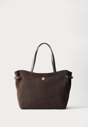 Τσάντα tote από σκούρο καφέ σουέντ με μαύρες λαβές και μικρό χρυσό λογότυπο μπροστά, τοποθετημένη σε απλό ανοιχτό φόντο.