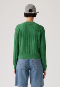 Personne portant un pull vert en maille torsadée, un short en denim bleu et une casquette à carreaux noir et blanc, debout avec le dos tourné à la caméra.