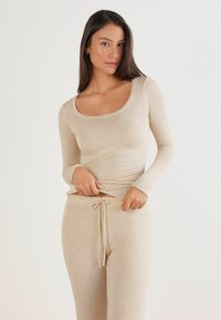 Beige Langarmoberteil aus weichem Material mit Rundhalsausschnitt, kombiniert mit passenden beigen Jogginghosen mit Kordelzug. Glatte Textur, figurbetontes Design.