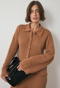 Knitted cardigan in warm brown with a collar, button closure, and textured pattern. Held with a black suede handbag featuring curved handles.