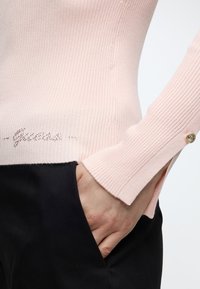 Gros plan d'une personne portant un pull à côtes rose clair avec un poignet fendu et un bouton doré, main dans la poche d'un pantalon noir.