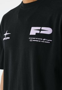 Camiseta de algodón negra de manga corta. Presenta texto gráfico y logotipos en púrpura en el pecho. Textura suave con un diseño de cuello redondo.