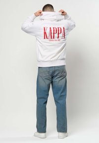 Mees kannab valget Kappa kapuutsiga dressipluusi, millel on punane logo, ja siniseid teksaseid, reguleerides kapuutsi, seisab seljaga palja heleda tausta ees.