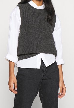 Personne portant un pull sans manches en tricot gris foncé sur une chemise blanche à boutons avec manches retroussées et un jean noir.