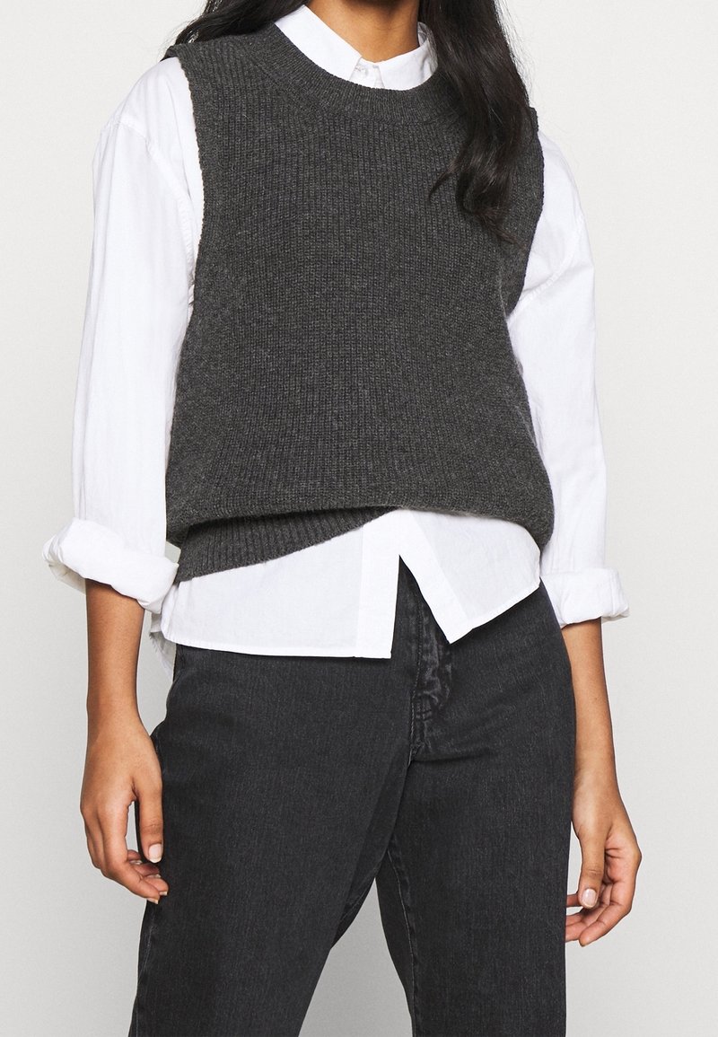 Personne portant un pull sans manches en tricot gris foncé sur une chemise blanche à boutons avec manches retroussées et un jean noir.