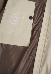 Beige ytterkläder med ett vattenavvisande och andningsbart foder. Har en svart tryckknapp och en vit teknisk informationsetikett inuti.