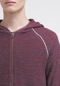 Mörkröd zip-up hoodie i strukturerad tyg. Har en huva med dragsko och kontrasterande sömmar vid axlarna. Dragkedja framtill.