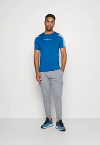 Kratke rokave modra športna majica z svetlo modrimi detajli, v kombinaciji s sivimi trenirkami. Ima logotip na prsih in lahek material.