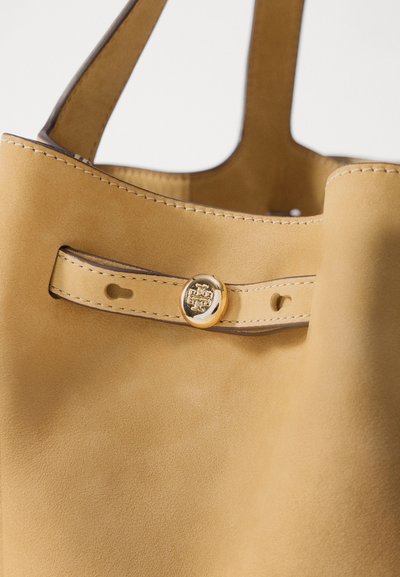 Gele suede schoudertas met een gladde textuur, voorzien van een gouden ronde knoopsluiting en contrasterende stiksels langs de randen.