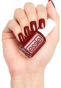 Flacon de vernis à ongles Essie tenu dans une main, présentant une couleur rouge profond brillante avec un capuchon blanc, mettant en valeur des ongles lisses et bien manucurés.