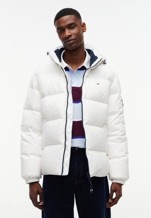 Mann trägt eine weiße Steppjacke über einem weißen, blau- und bordeauxfarben gestreiften Poloshirt mit dunklen Hosen, steht vor einfachem Hintergrund.