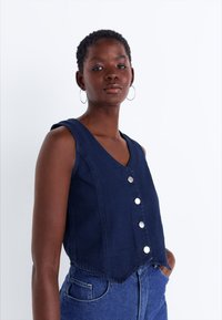 Λ navy blue τζιν γιλέκο με V-neckline, μπροστινές κουμπώματα και κοντή σχεδίαση. Διαθέτει χωρίς μανίκια κοπή και ξέφτια στη βάση για επιπλέον υφή.