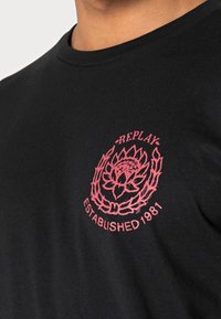 Czarny t-shirt z różowym nadrukiem na klatce piersiowej, przedstawiającym wzór kwiatowy, wieniec laurowy oraz napis „REPLAY ESTABLISHED 1981.”