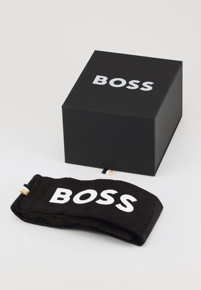 Un bandeau en tissu noir avec le logo "BOSS" en blanc, posé à côté d'une boîte rectangulaire noire arborant le même logo en blanc.