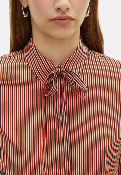 Vrouw in een verticaal gestreepte blouse met rode, marineblauwe en beige strepen, met een kleine geknoopte strik bij de kraag en zilveren ringoorbellen.