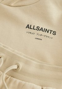 Naturalna bawełniana tkanina w jasnym beżu, z okrągłym dekoltem i talią regulowaną sznurkiem. Czarne nadrukowane logo "ALLSAINTS" i "LONDON."