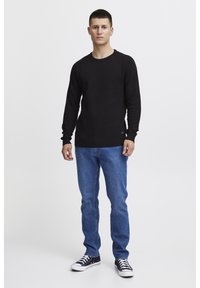 Cămașă neagră texturată cu mânecă lungă, de tip crew neck, combinată cu blugi albaștri și pantofi sport negri. Design simplu, fără modele sau logo-uri vizibile.