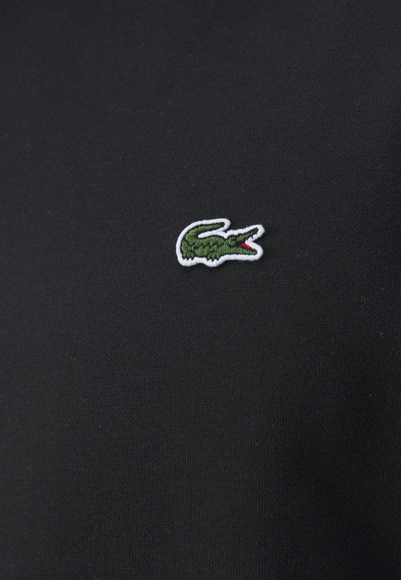 Schwarzes Baumwollpolo-Shirt mit einem kleinen, gestickten grünen Krokodil-Logo mit roten Akzenten auf der linken Seite der Brust.