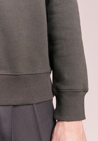 Sudadera verde oscura con puños y dobladillo acanalados, que presenta una tela suave de textura. Los detalles de costura visibles realzan el diseño.