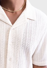 Short-sleeve white knitted shirt featuring vertical stripes, button front, and textured fabric with small open patterns, complemented by a gold chain necklace.