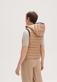 Kvinna sedd bakifrån, iklädd en beige, vadderad ärmlös väst med huva, en kortärmad ribbad topp och beiga byxor.