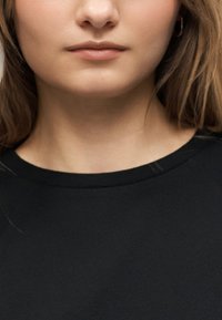 Κοντινό πλάνο του κάτω μέρους του προσώπου και του λαιμού μιας γυναίκας με φυσικά χείλη και ανοιχτόχρωμο δέρμα, που φοράει μαύρο μπλουζάκι με λαιμόκοψη σε σχήμα κύκλου.