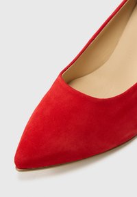 Sarkanas zamšādas kurpes ar smailu purngalu un gludu tekstūru, ar zemu izgriezumu priekšpusē un nedaudz izliektu aizmuguri. Zeltainas krāsas zolīte detaļa.