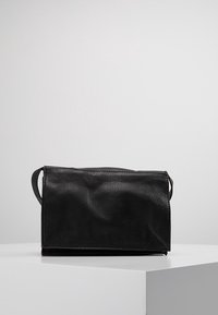 Borsa in pelle nera con superficie testurizzata, forma rettangolare, due manici laterali e chiusura a pattina, posizionata su un'esposizione minimalista.