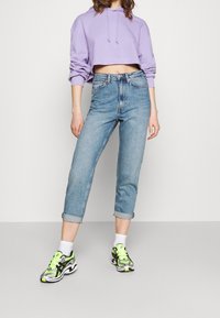 Felpa corta con cappuccio viola, jeans azzurri a vita alta con risvolti e sneakers nere e verde neon con dettagli grigi.