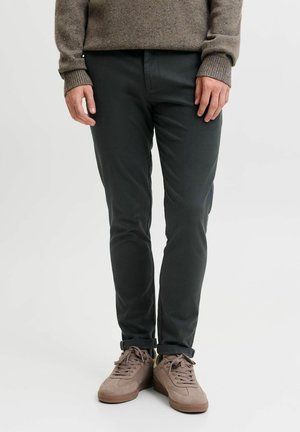 Muž nosi tmavé slim fit nohavice s vyhrnutými lemy, béžové tenisky a hnedý pletený sveter, stojaci pred jednoduchým svetlým pozadím.