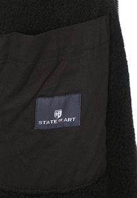 Zwarte jassenvoering met een getextureerd fleece-materiaal. Inclusief een zwart stoffen label met het "STATE OF ART" logo en contrasterende accenten.