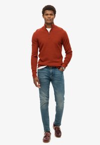 Suéter de cuello con cremallera color óxido, jeans de mezclilla claros y zapatos rojo oscuro. El suéter presenta un acabado texturizado y un diseño ajustado.